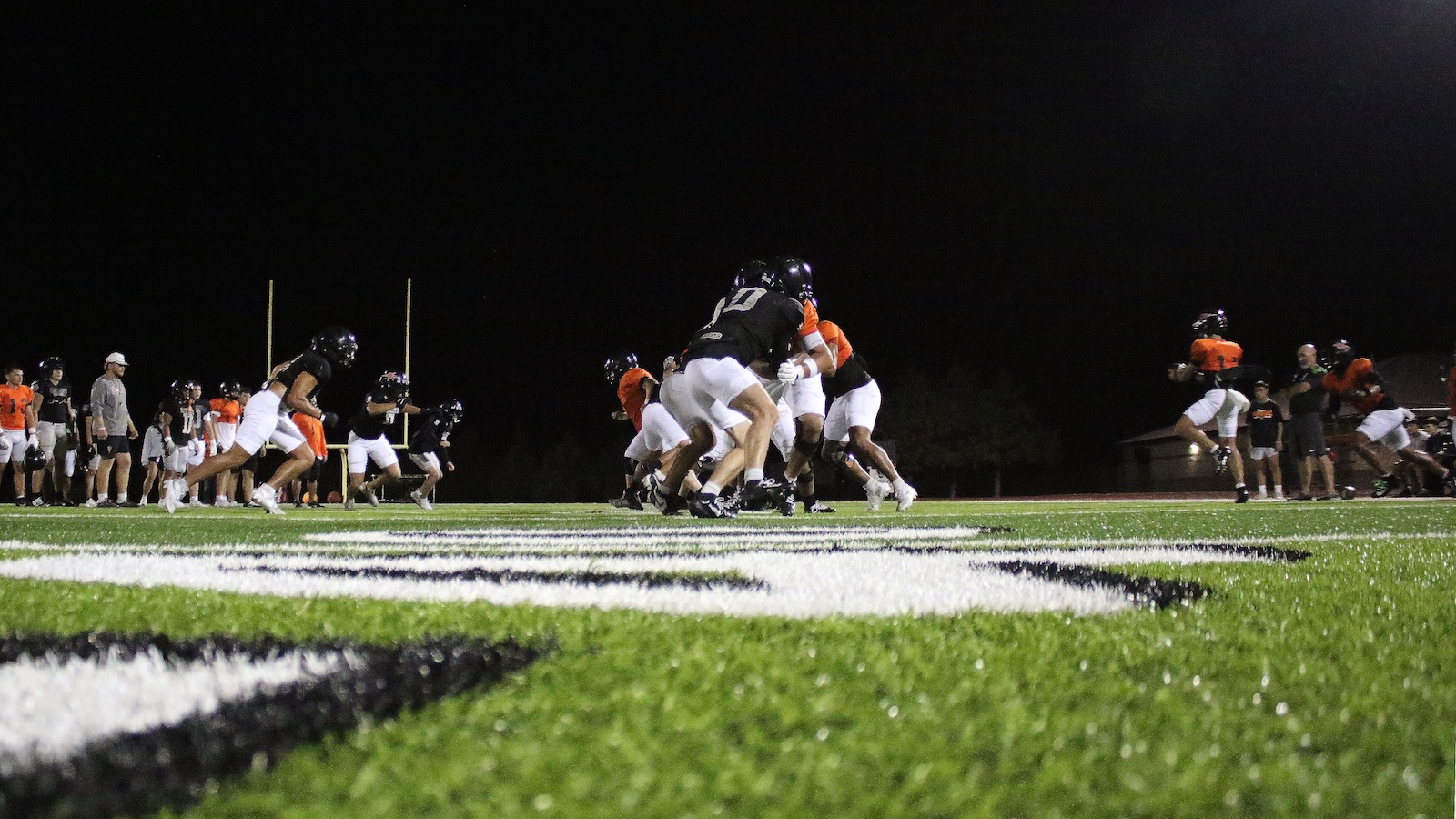 Nighttime football game with players in black and orange colliding at the line of scrimmage under bright stadium lights on a green field.