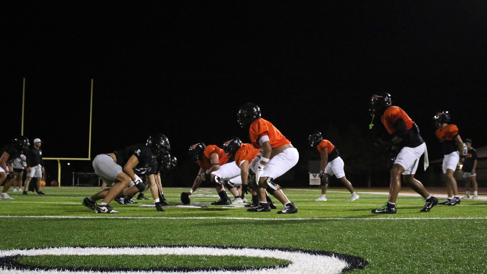 Nighttime football practice with players in orange jerseys lined up at the line of scrimmage on a green synthetic turf field.