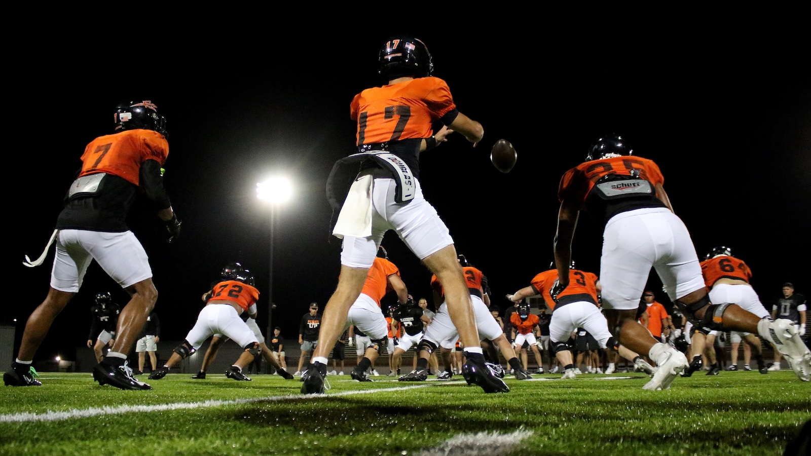 Football players in orange jerseys practice on a night-lit field, with a quarterback preparing to throw.
