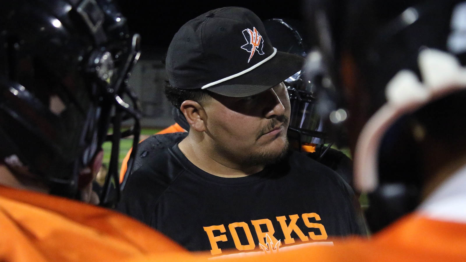 Close-up of a man in a black cap and black shirt reading 'FORKS', surrounded by players in orange uniforms during a team huddle.