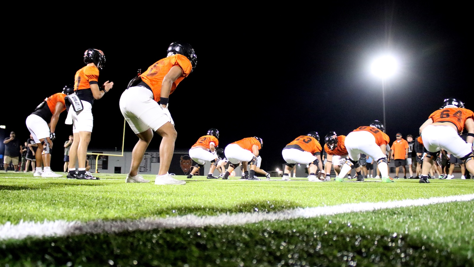 Football players in orange jerseys crouch at the line during a nighttime practice on a lit football field.