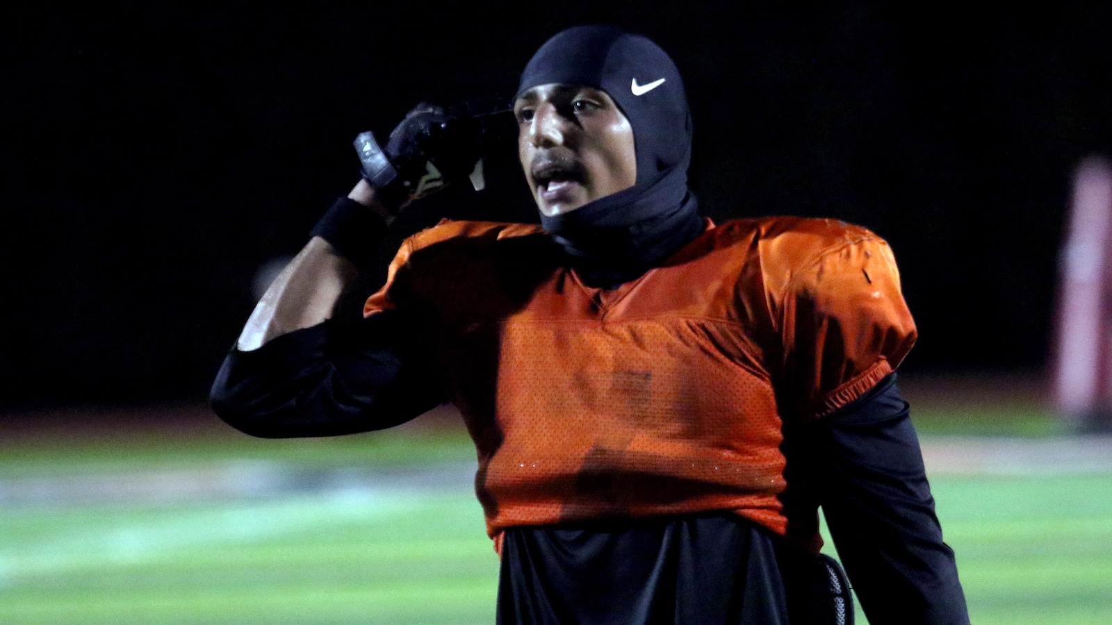 Football player in an orange practice jersey, wearing a black undershirt and helmet, on a night field, hand to ear as he calls out.