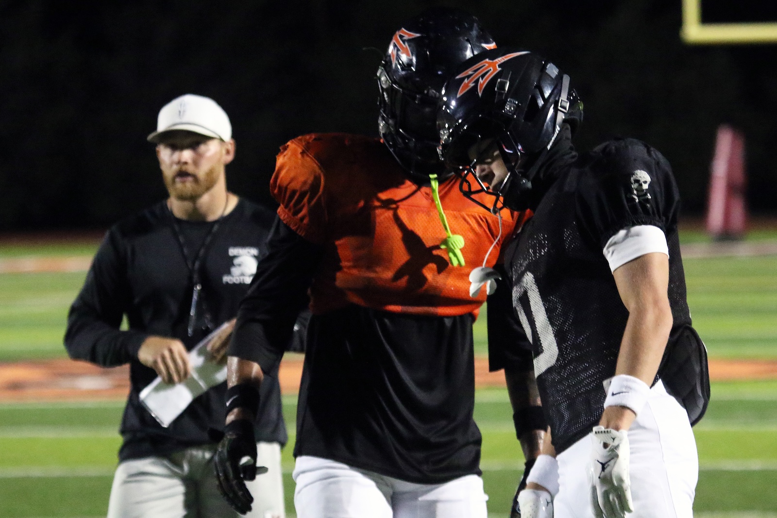 Two football players wearing black uniforms and helmets lean together on a night practice field while a coach in a white cap watches nearby.