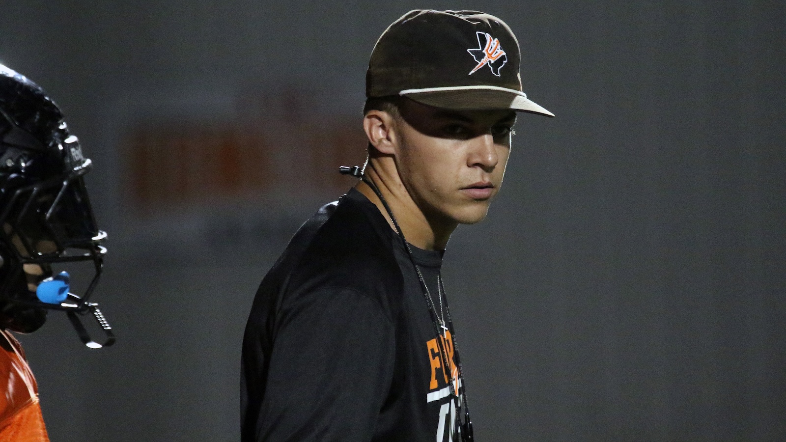 Young man in a black cap and dark shirt looks to the side during a sports practice or game, with a headset-wired mic on a teammate visible at left.
