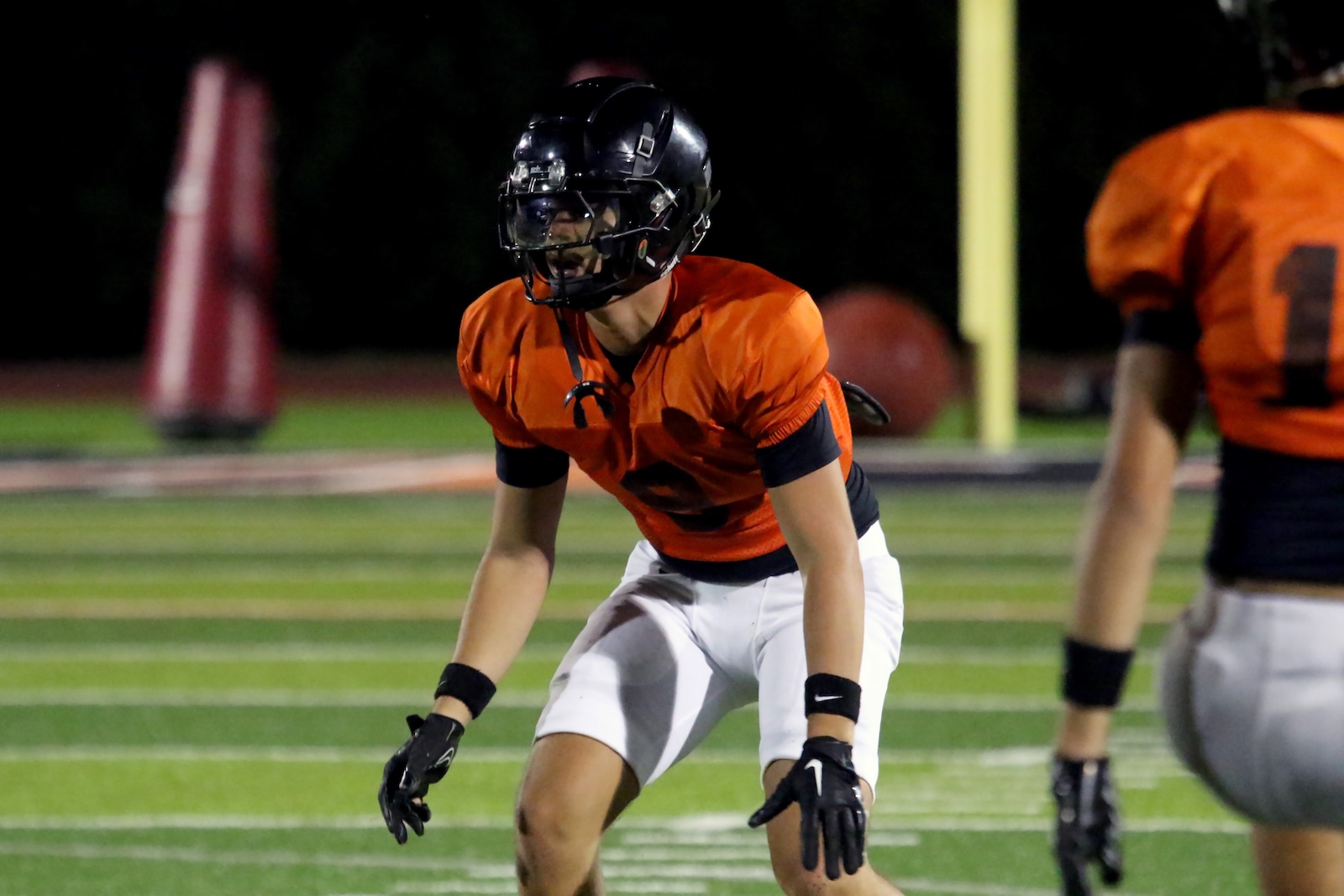 Young football player in an orange jersey and white pants crouches in a ready stance on a nighttime field, wearing a black helmet and gloves on both hands.