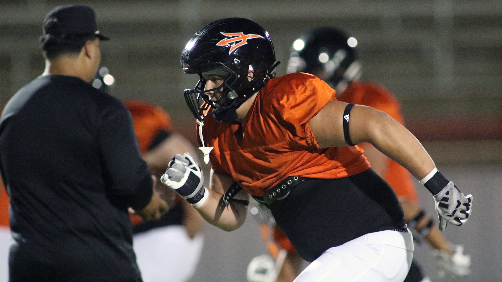 American football practice: a player in an orange jersey and black helmet sprints toward a coach on the field at night.