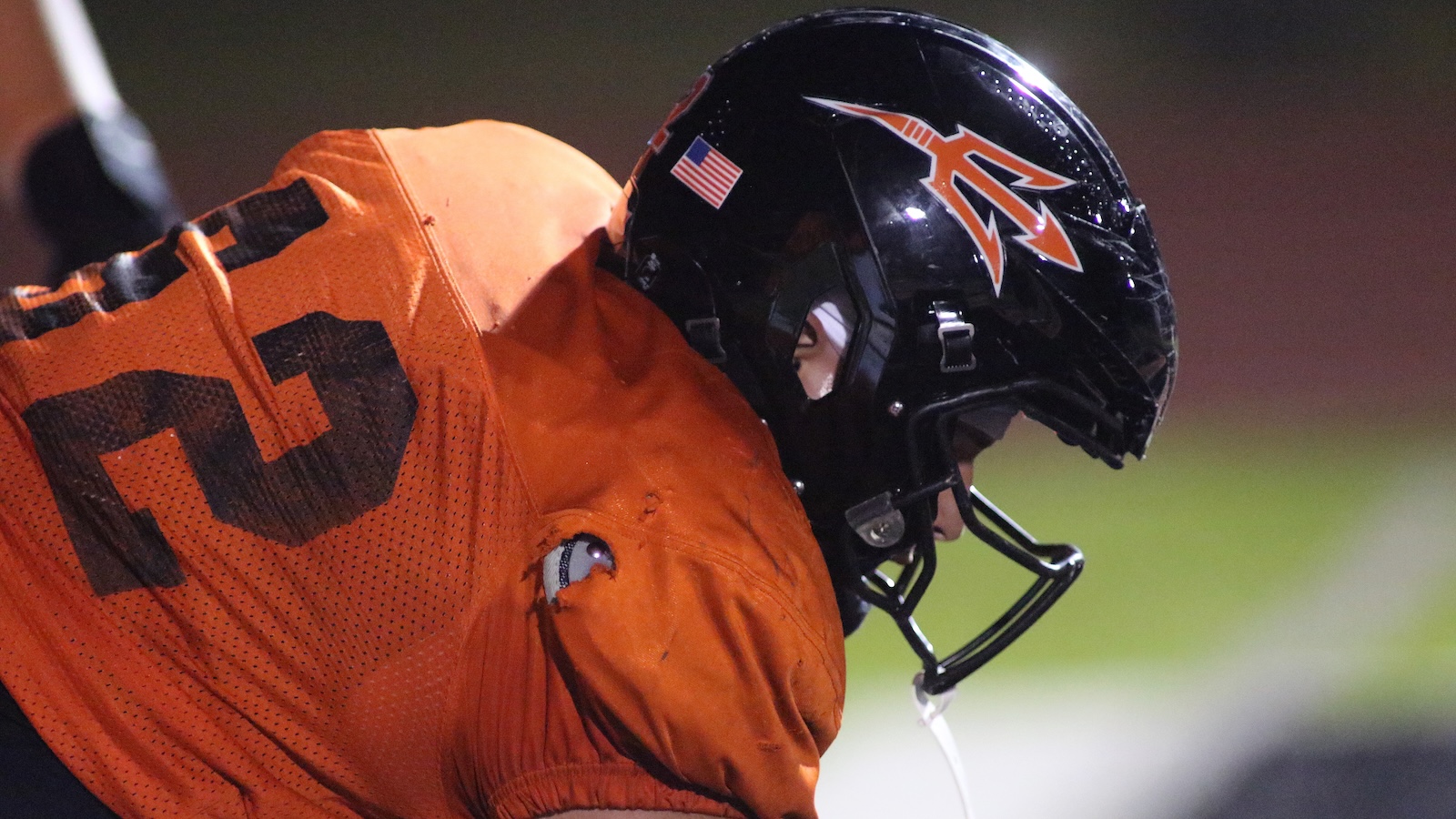 American football player wearing an orange jersey and a black helmet leaning forward on the field during play. Includes a small American flag sticker on the helmet and an orange logo on the side along with a faceguard.