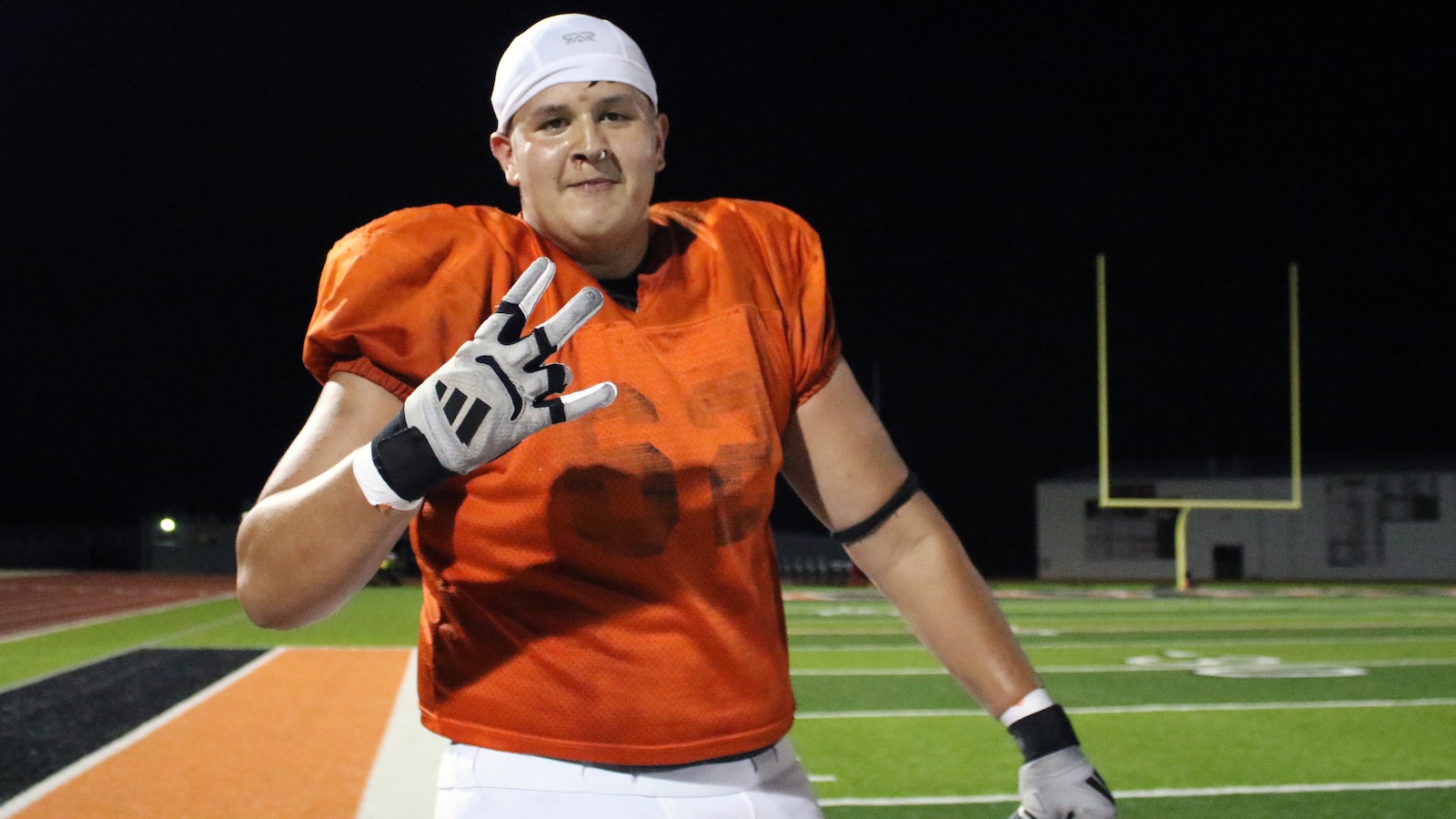 Football player in an orange jersey and white cap on a night field, reaching out with a gloved hand as a gesture of a high five.