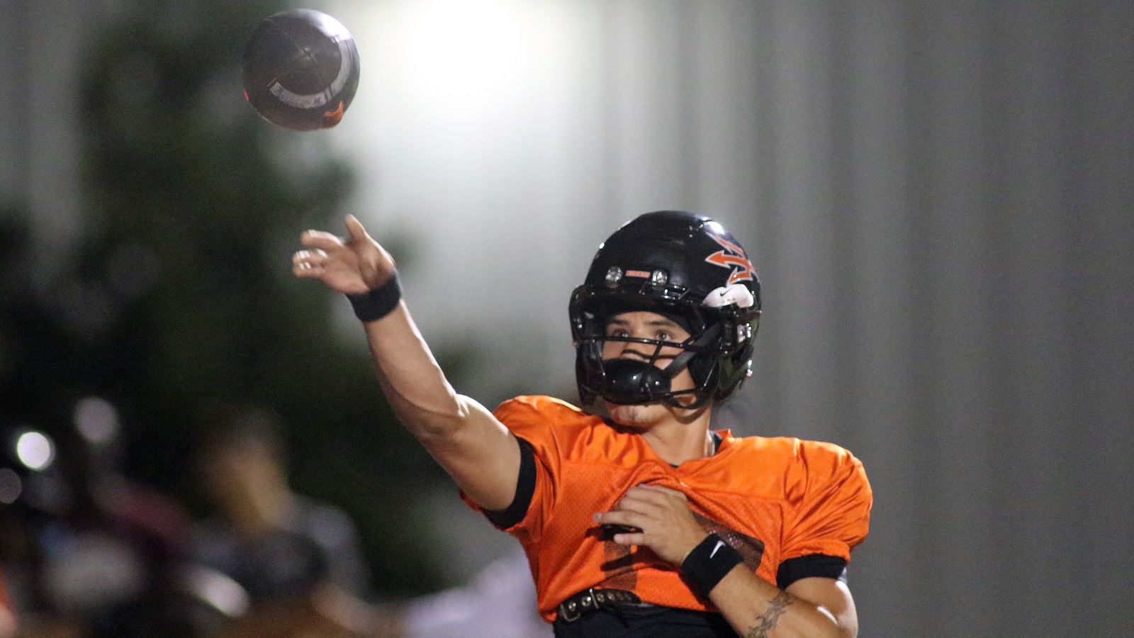 Football quarterback in orange jersey throws a ball mid‑air, black helmet on, stadium lights in background