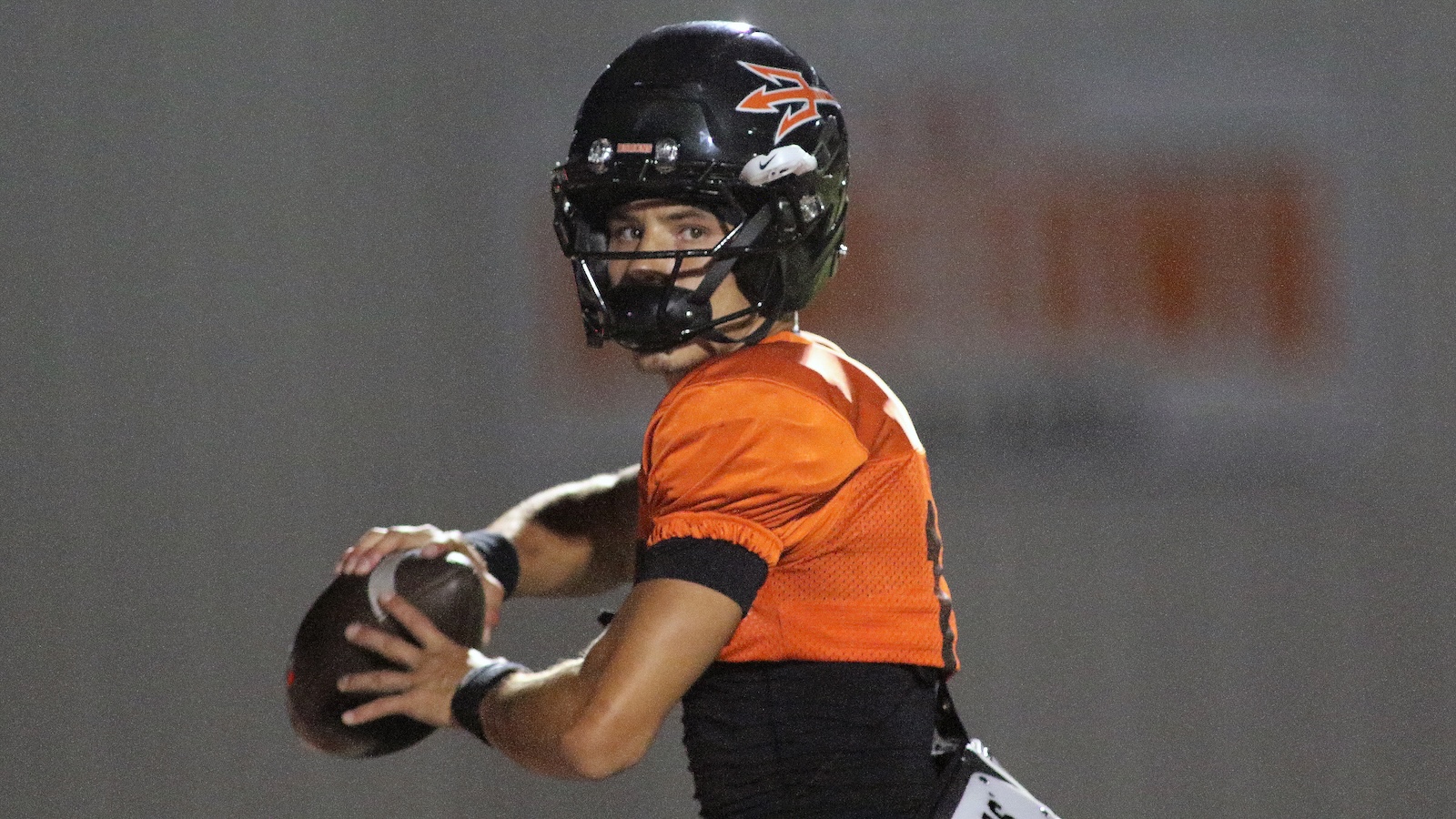 Quarterback in orange jersey and black helmet holding football, looking to pass during a nighttime game