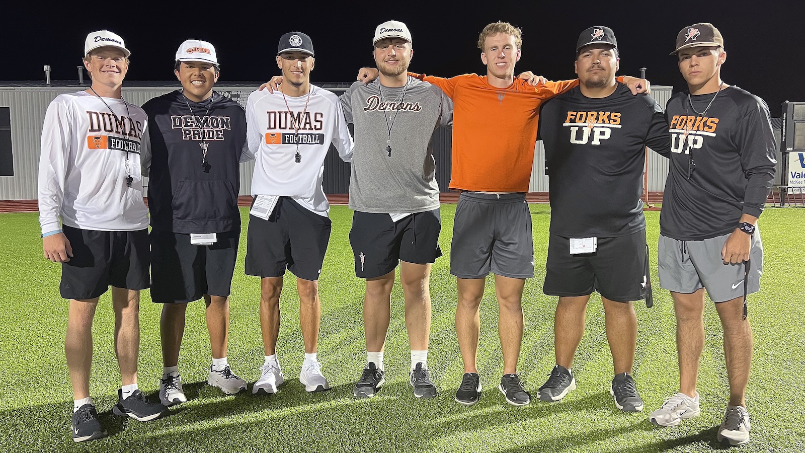 Dumas - Grow Your Own | Press Pass Sports Eight young men stand shoulder to shoulder on a lit football field at night, arms around each other, wearing team shirts and hats for a group photo.