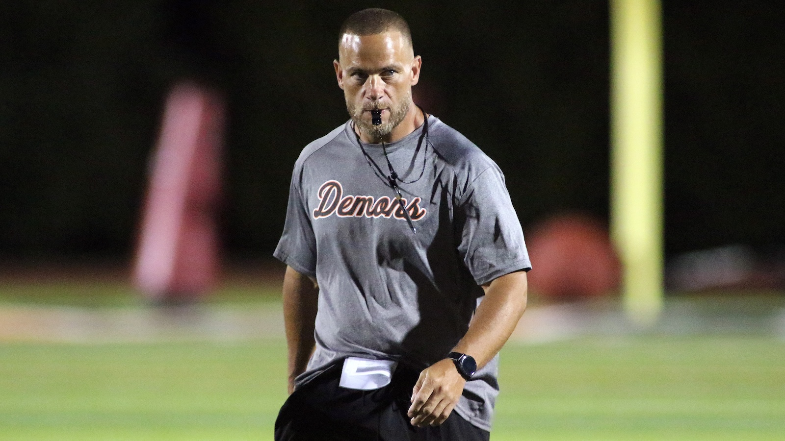 Male coach on a nighttime sports field, gray 'Demons' shirt, whistle in mouth, watch on wrist, focused forward