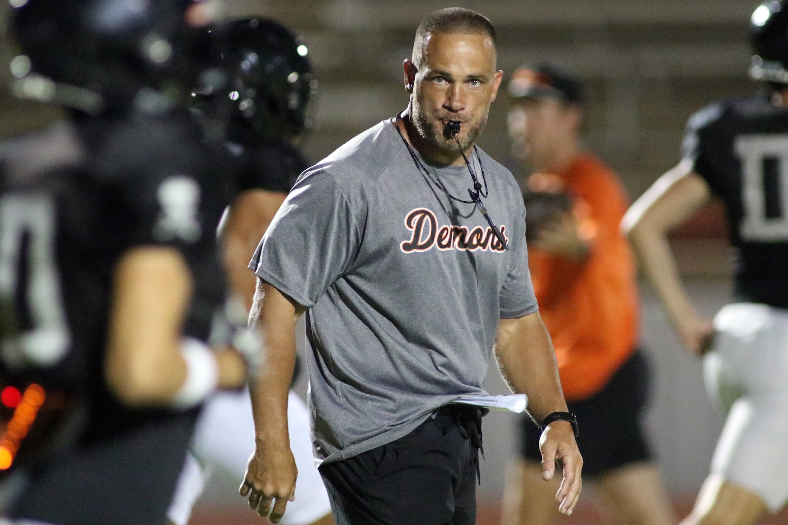 Male football coach in a gray Demons shirt with whistle in mouth, standing on the sideline during practice.