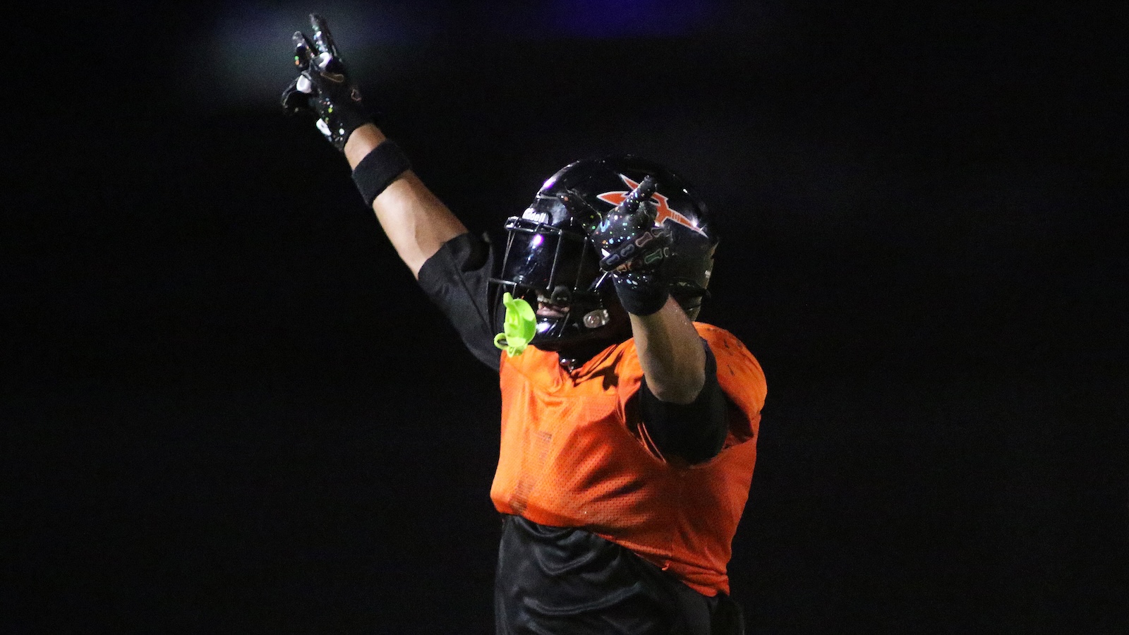 Football player celebrating with arms raised at night, wearing an orange jersey and black helmet with gloves.