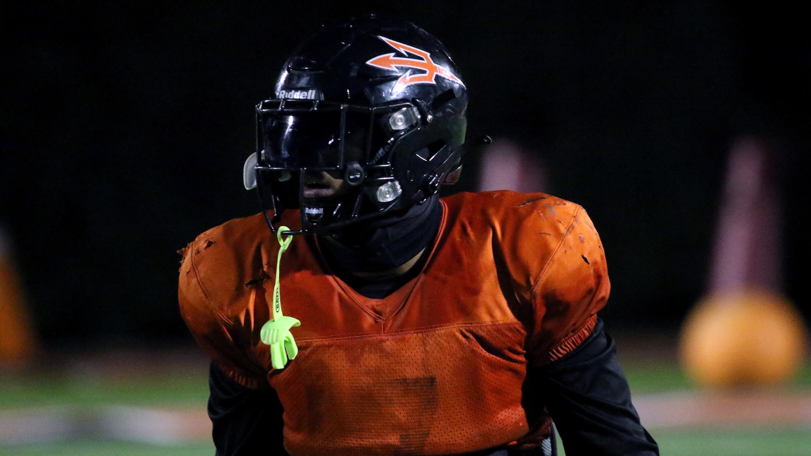 Football player in orange jersey and black helmet during a night practice on a dim field.