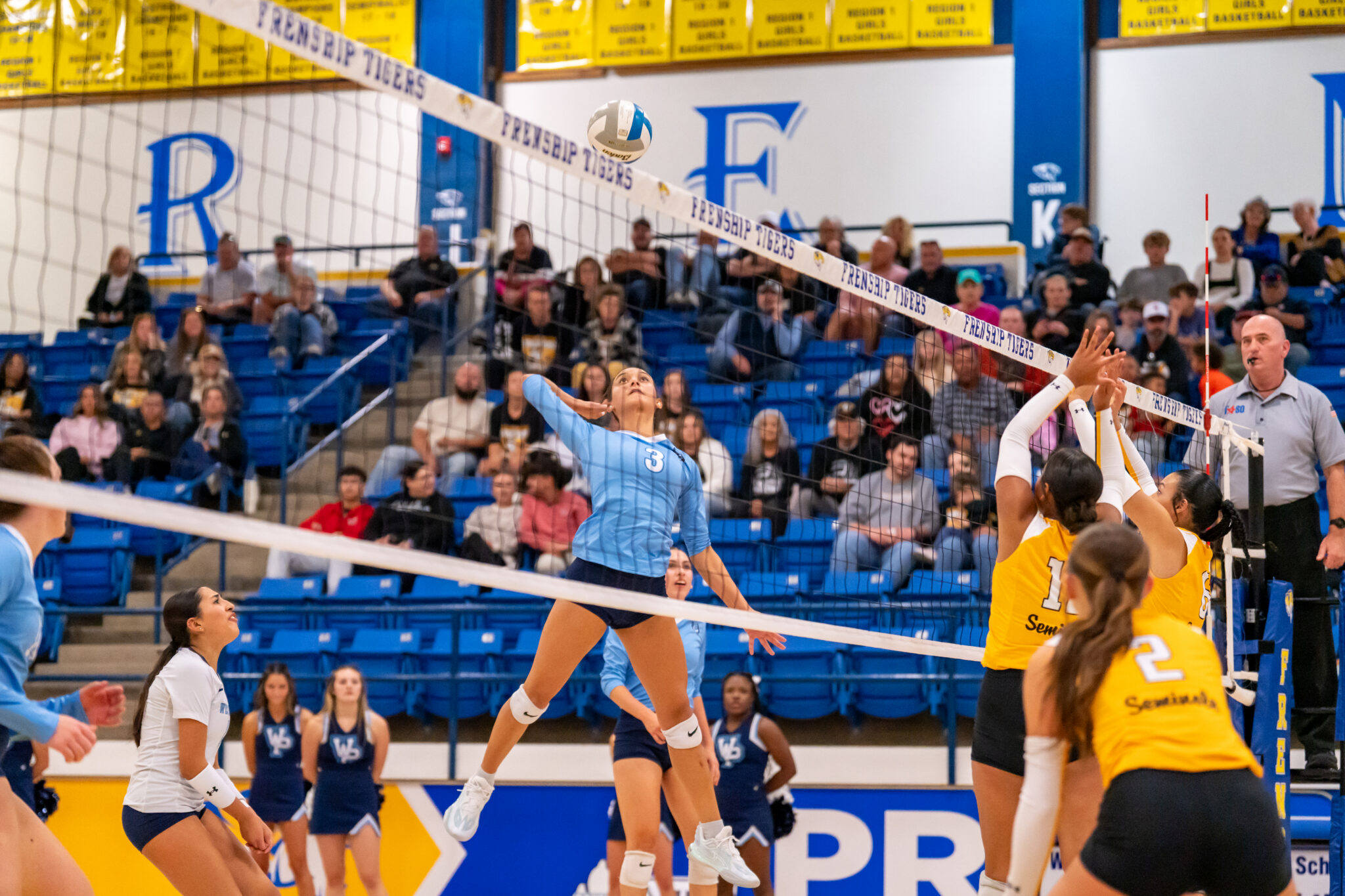 DSC01990 | Press Pass Sports Female volleyball player in a light blue uniform jumps to spike the ball over the net as teammates and spectators watch in a crowded gym.