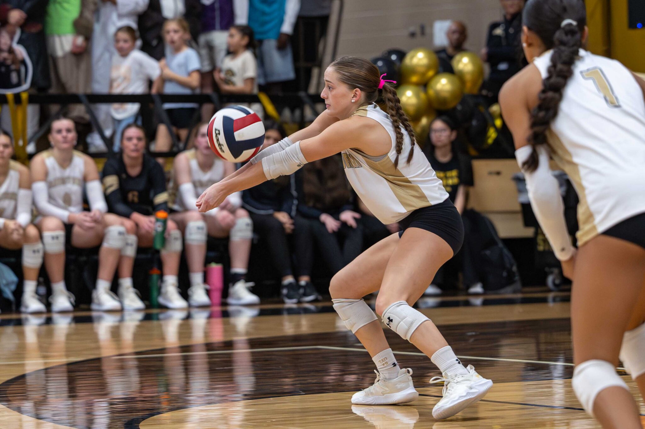 THS-AHS-VB-22 | Press Pass Sports Female volleyball player in white and gold digs the ball with forearms extended during a match, crowd watching in the background.
