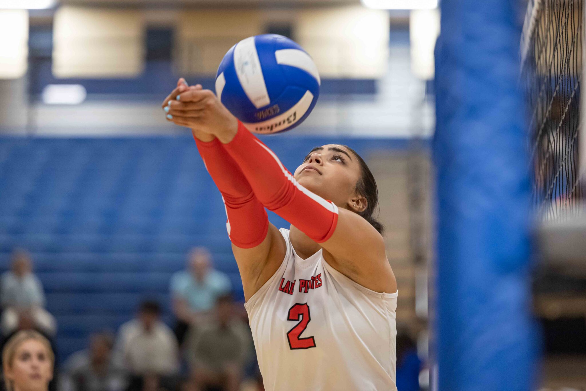 LCHS-PDHS-VB-12 | Press Pass Sports Female volleyball player in a white jersey with number 2 raises arms to block a blue volleyball near the net in a gym.