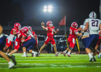 100A6242 | Press Pass Sports Quarterback in a red jersey throws a pass during a night football game as teammates block and defenders approach.
