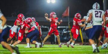 Quarterback in a red jersey throws a pass during a night football game as teammates block and defenders approach.