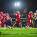 100A6242 | Press Pass Sports Quarterback in a red jersey throws a pass during a night football game as teammates block and defenders approach.