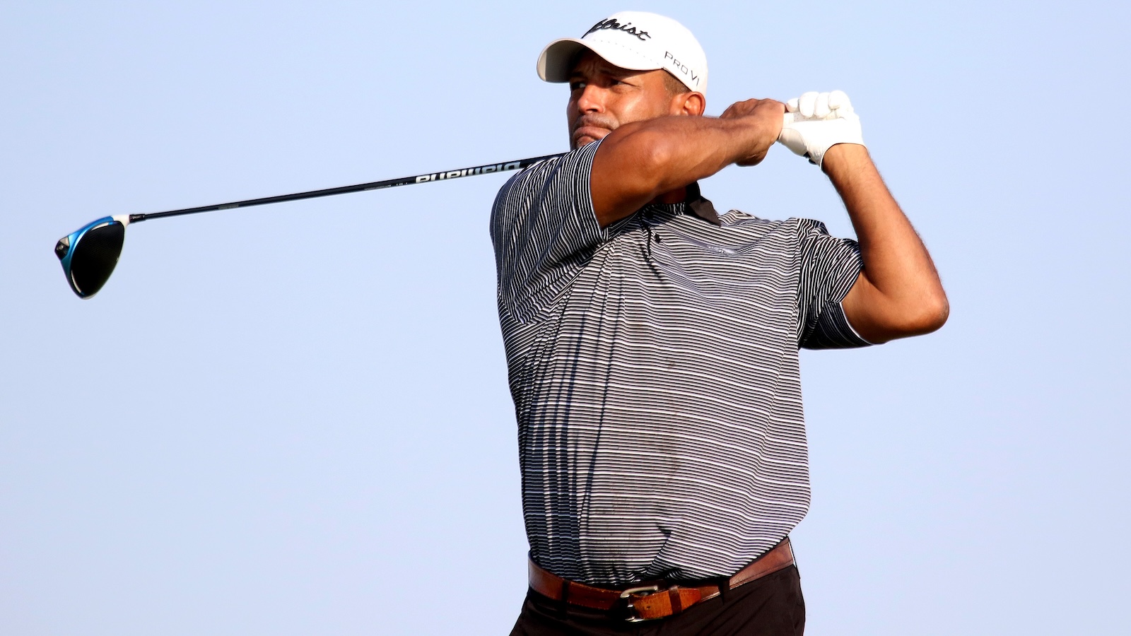 Male golfer in a striped polo and white cap swinging a driver against a clear blue sky.