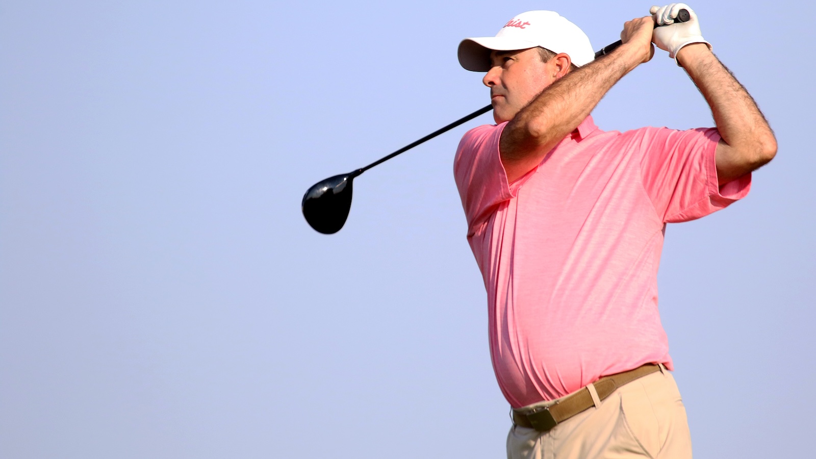 Golfer in a pink polo shirt and beige pants mid-swing with a driver against a clear blue sky.