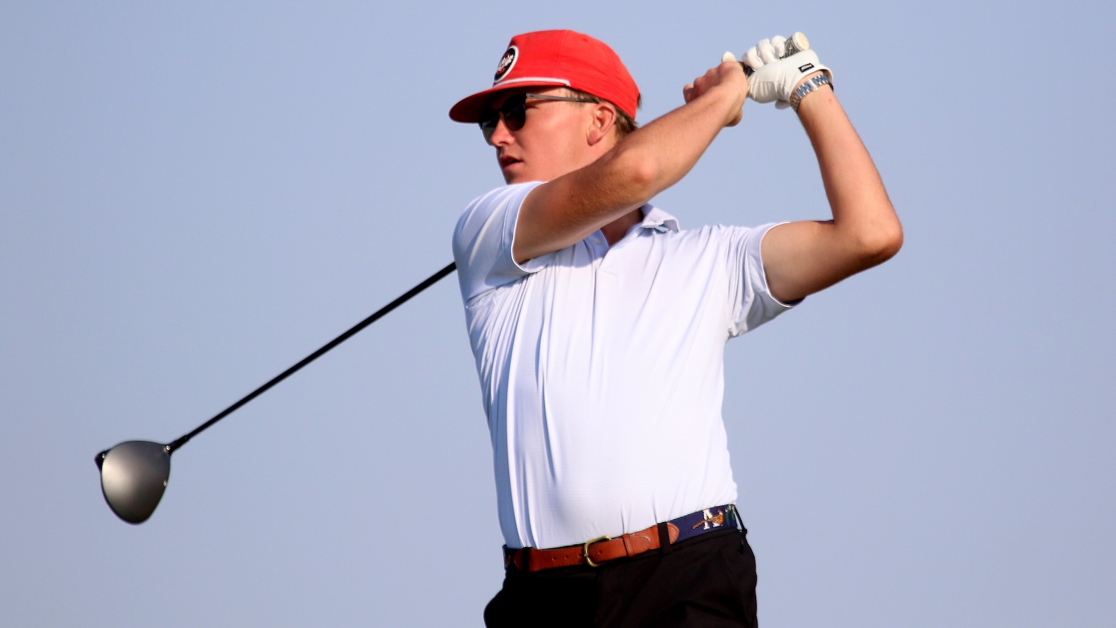 Male golfer in a white polo and red cap mid-swing, against a clear blue sky, wearing sunglasses.