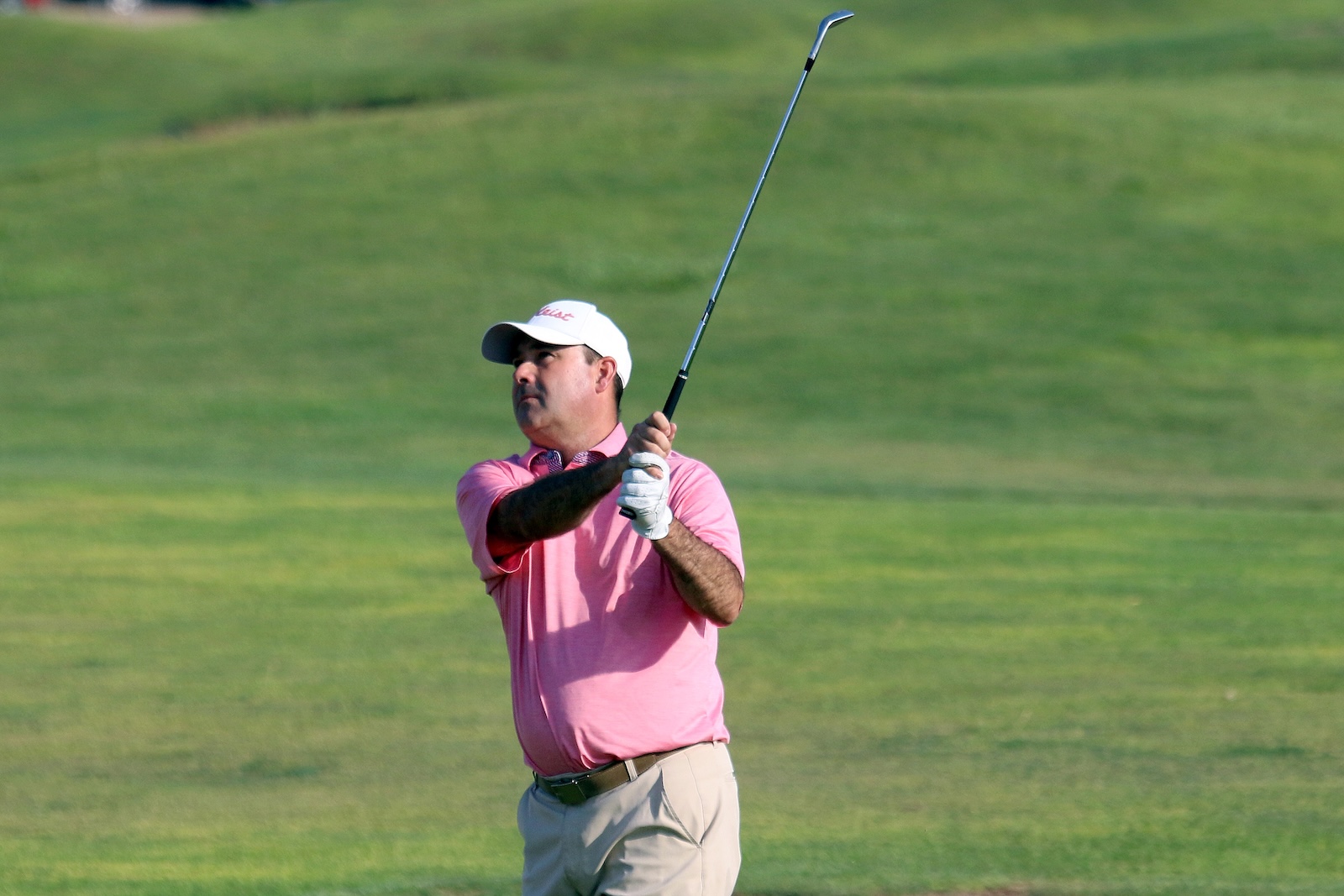 Male golfer in a pink polo shirt and white cap swinging a golf club on a green course