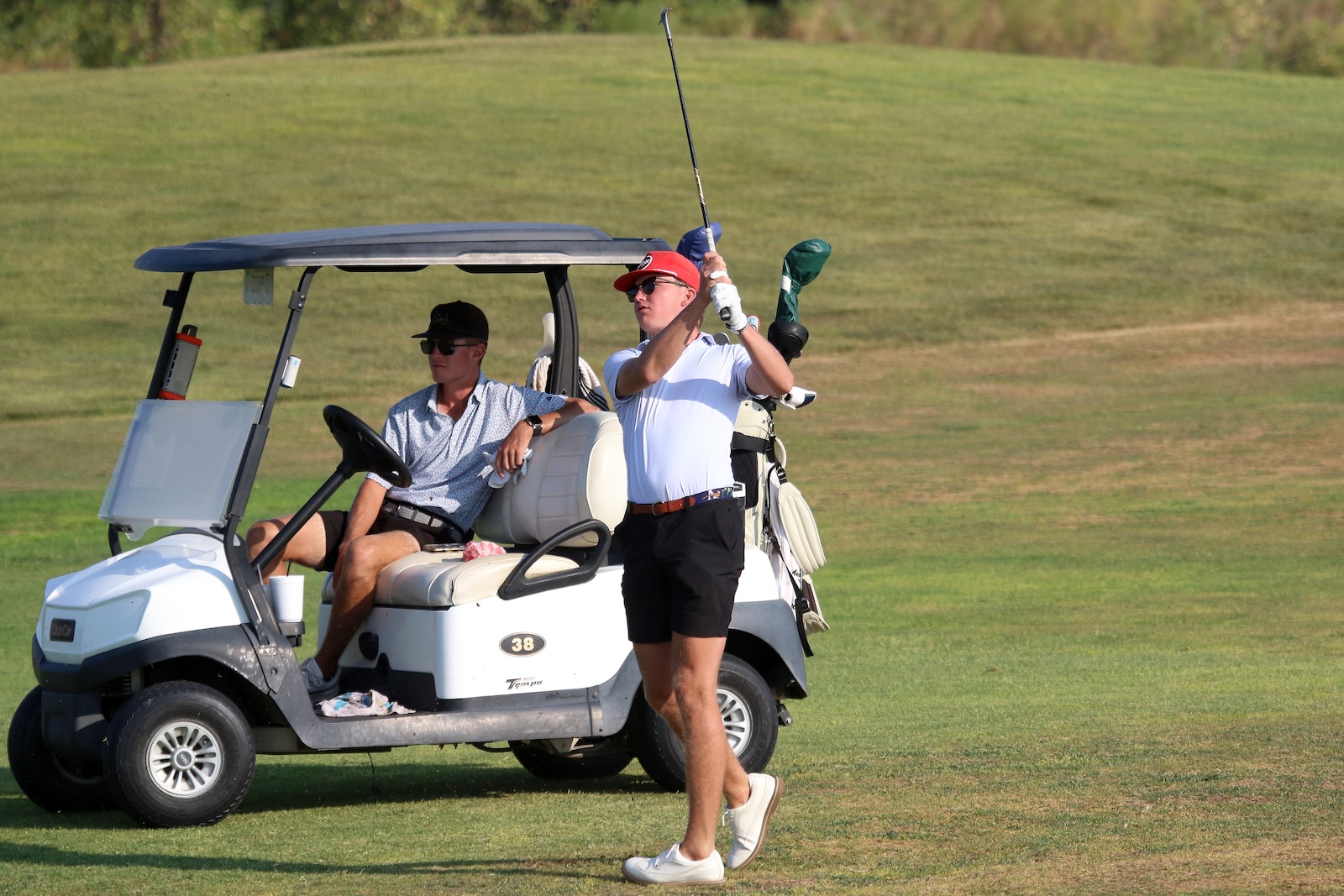 A man in a red cap swings a golf club beside a white golf cart on a grassy course, another man sits in the cart watching.