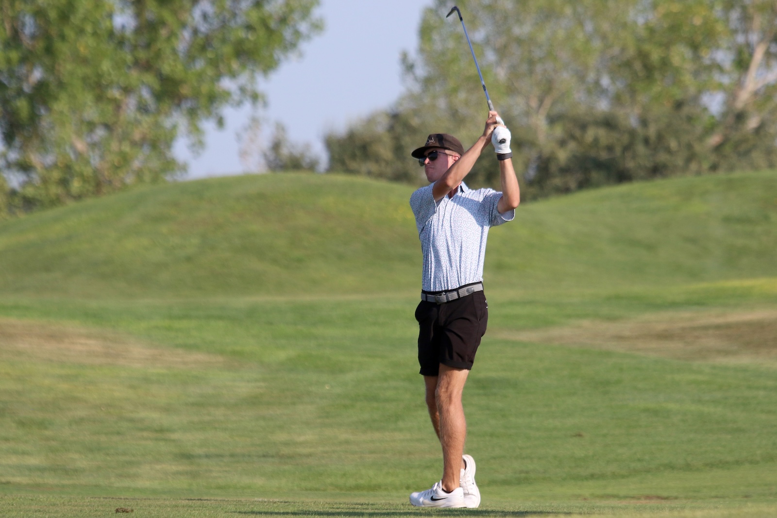 Male golfer mid-swing on a sunny golf course, wearing a patterned polo, black shorts, white shoes, and a cap with sunglasses.