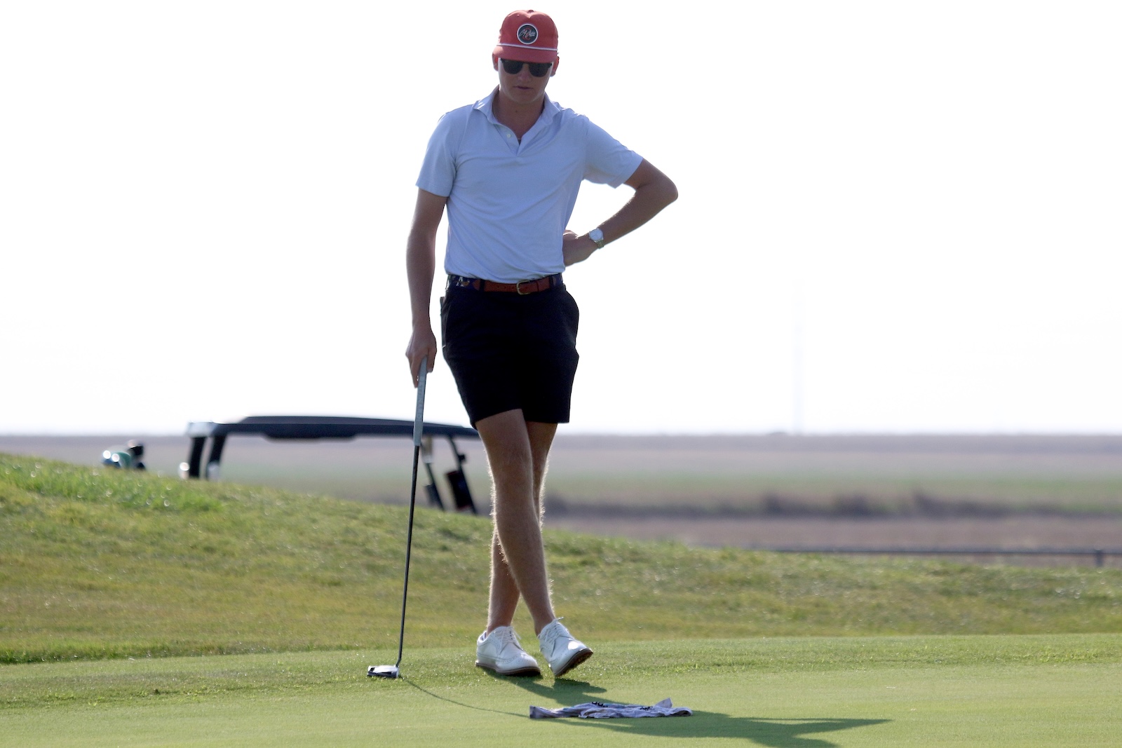 Man on a golf green preparing to putt, wearing a white polo, black shorts, white shoes, red cap and sunglasses while holding a putter.