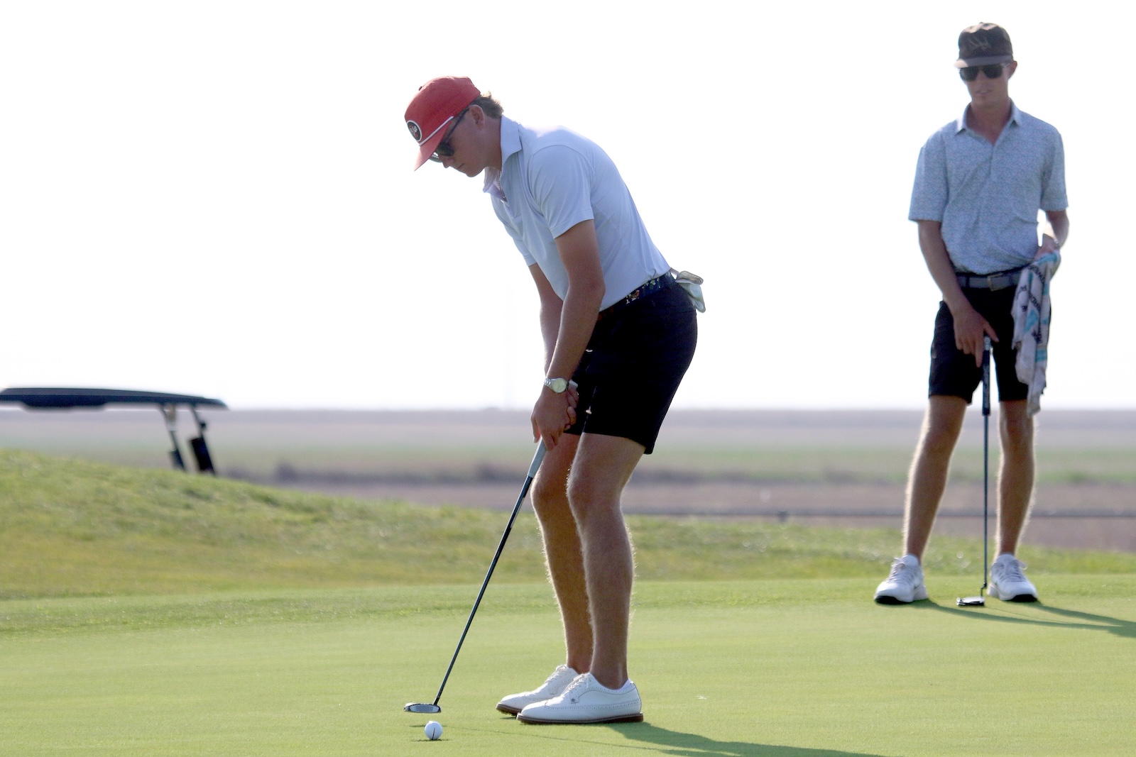 Two golfers on a green: one bends to putt the ball, the other stands nearby with a towel and club watching.