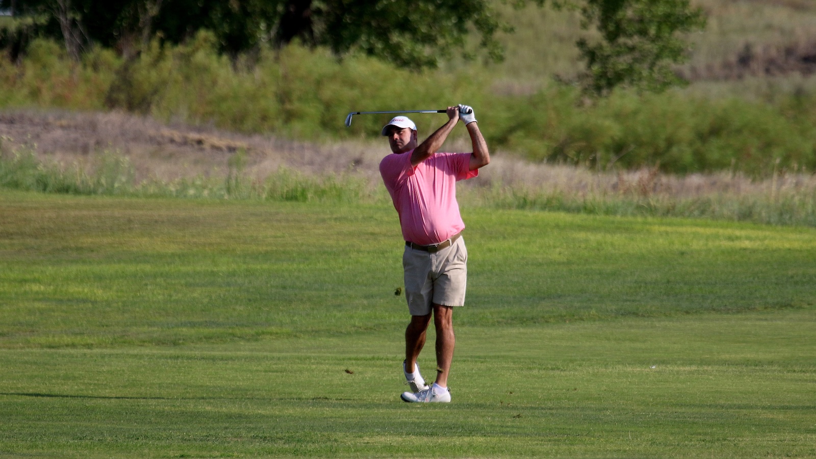 Male golfer in a pink polo and white cap mid-swing on a green golf course with trees in the background.