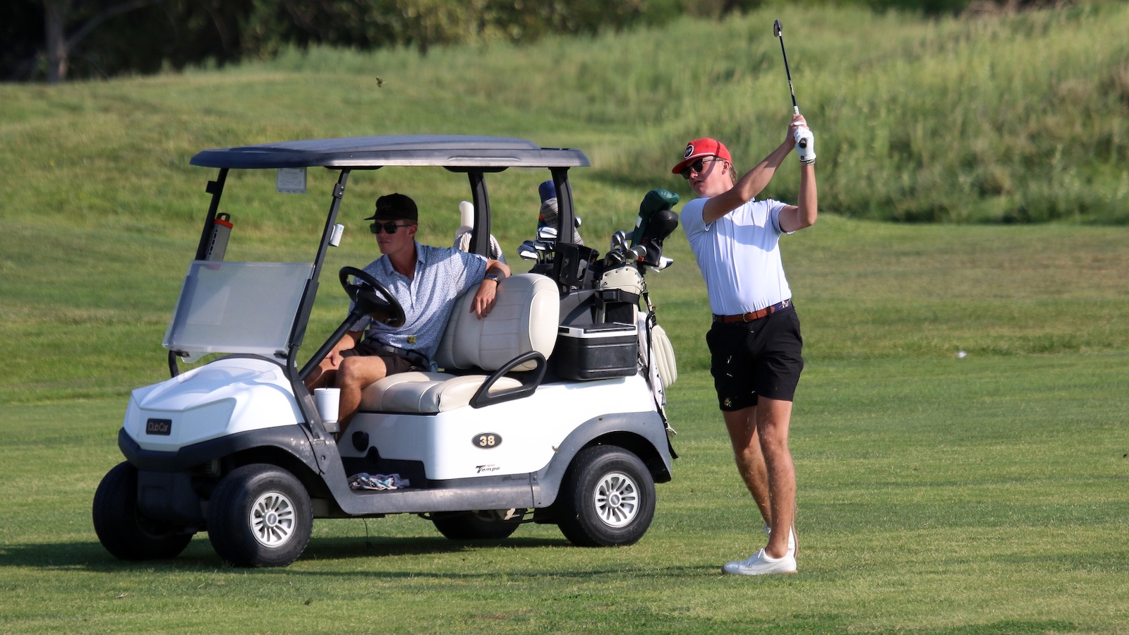 Two men on a golf course: one in a white polo swings a club while standing beside a parked golf cart, the other sits in the cart watching.