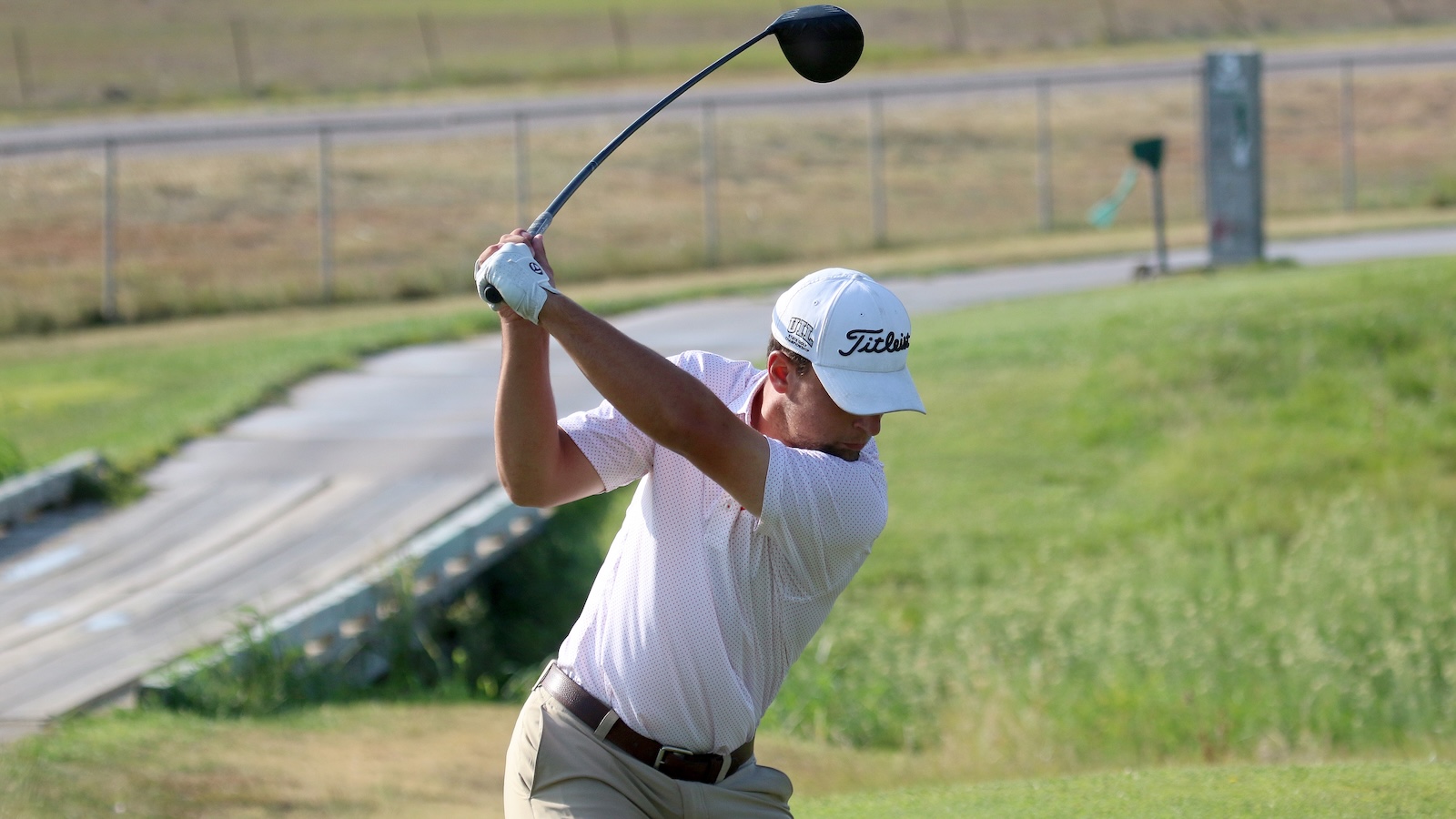 Golfer in white polo and cap swinging a driver on a grassy course with a fence in the background