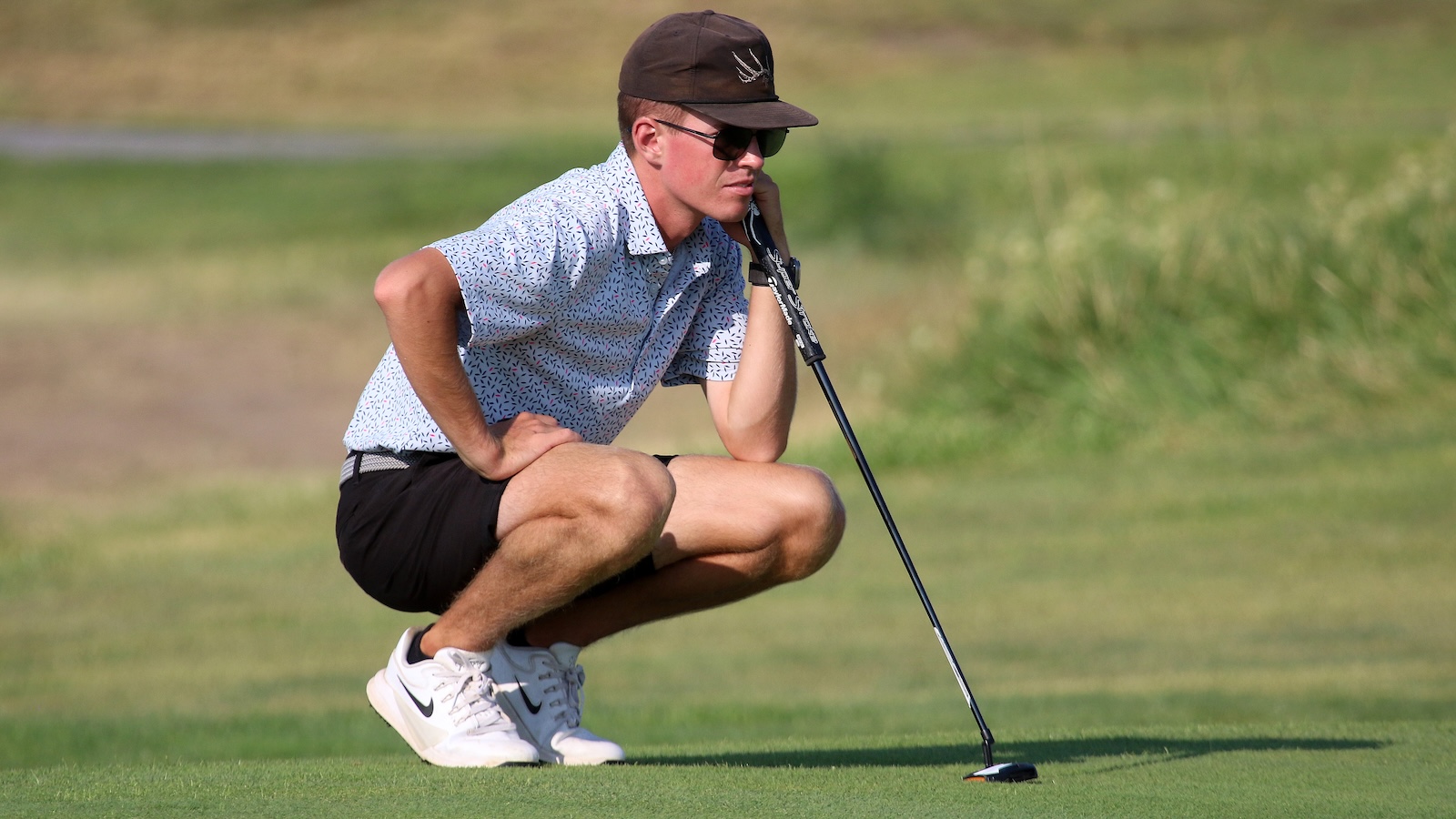 Golfer crouching on a putting green, lining up a putt with a putter, wearing sunglasses and a brown cap.