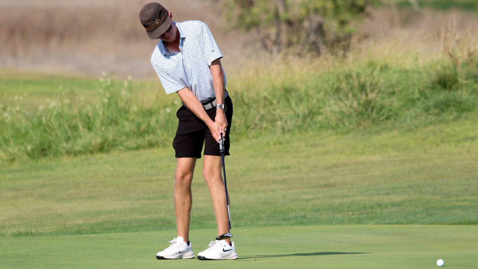 Man wearing a cap and patterned polo shirt lines up a putt on the green, holding a golf club over a short backswing.