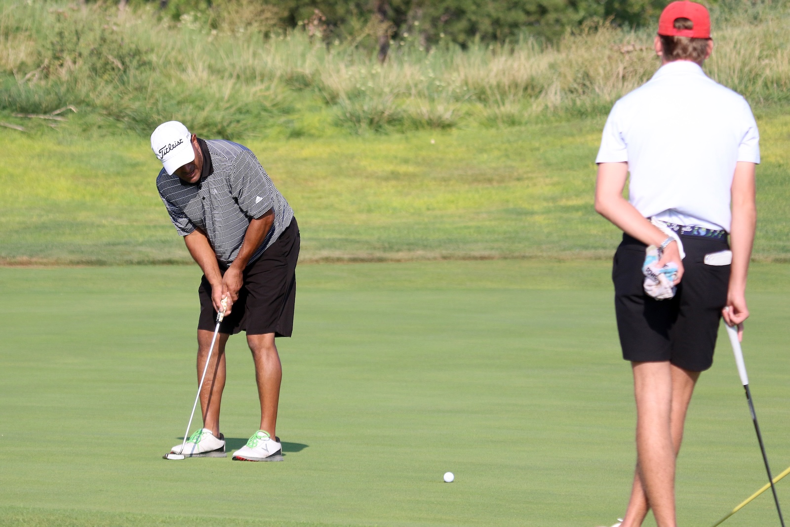 Golfer lining up a putt on a green while another player watches in the background.