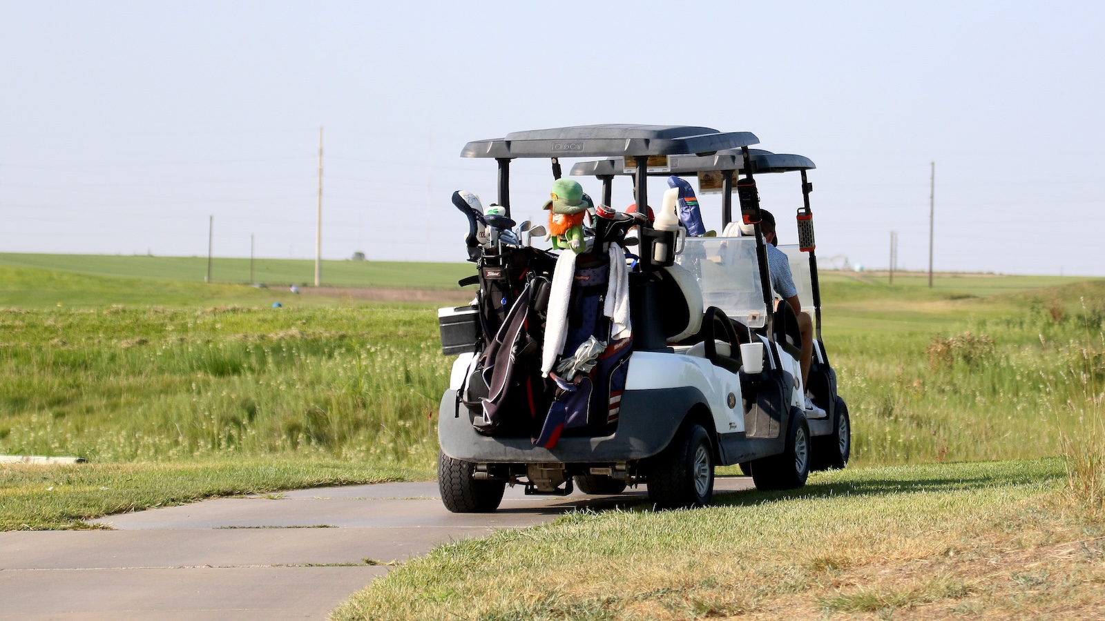 Golf cart driving along a cart path with multiple golf bags and clubs attached to the back on a grassy golf course.