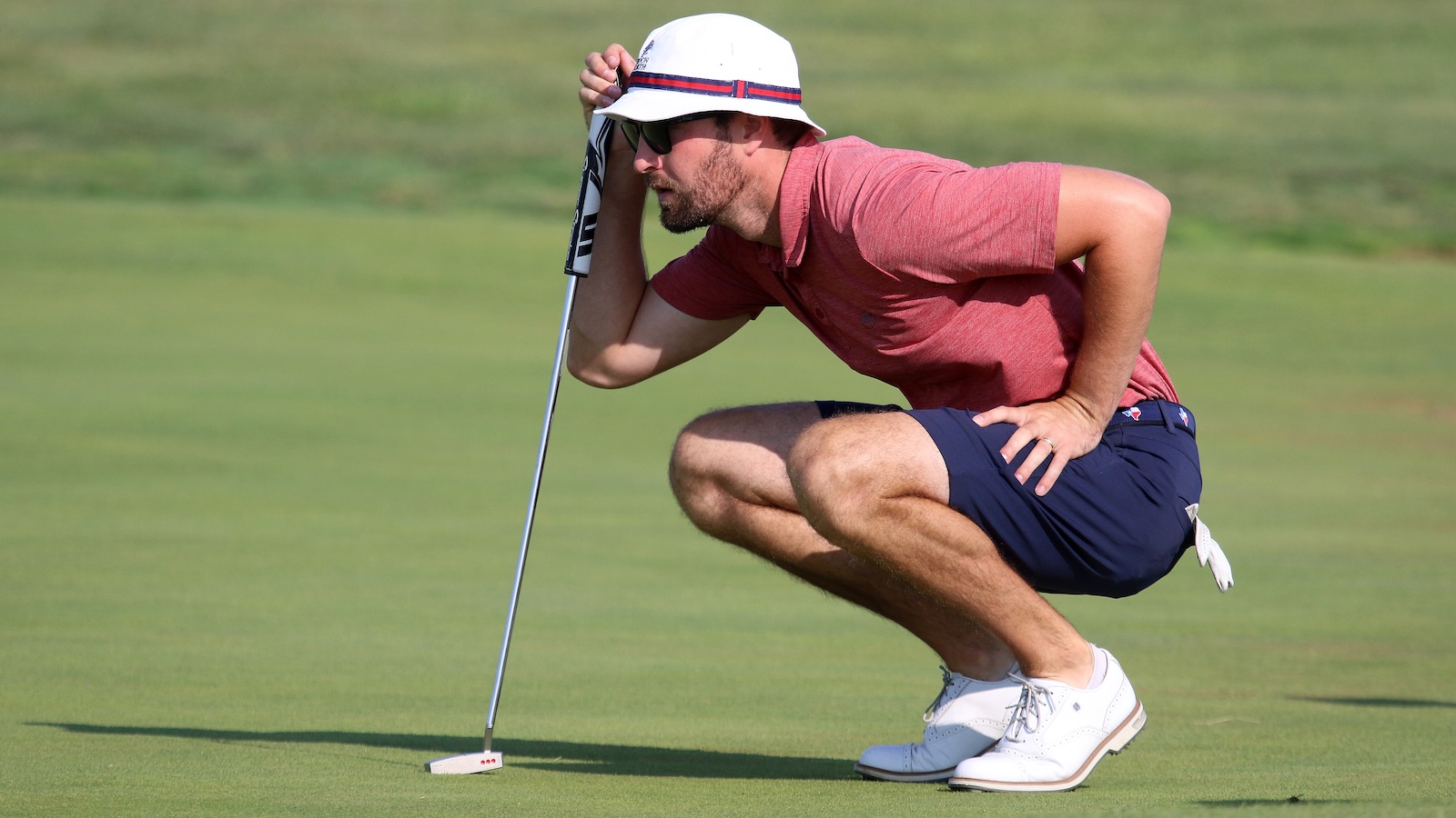 Golfer in a red shirt and white hat crouches on the green to line up a putt with a putter.