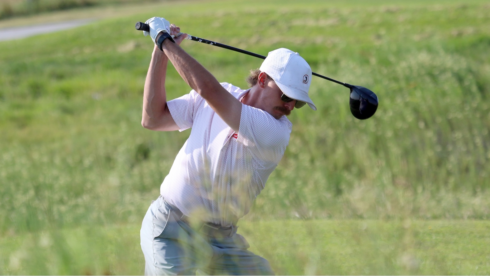 Man in a white polo and cap mid-back swing on a golf course, holding a driver with both hands