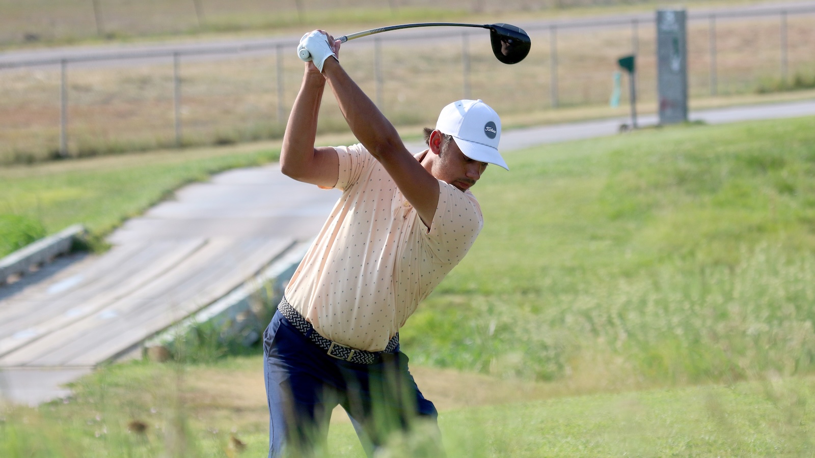 Male golfer in a pale polo and white cap mid-swing on a golf course with a driver.