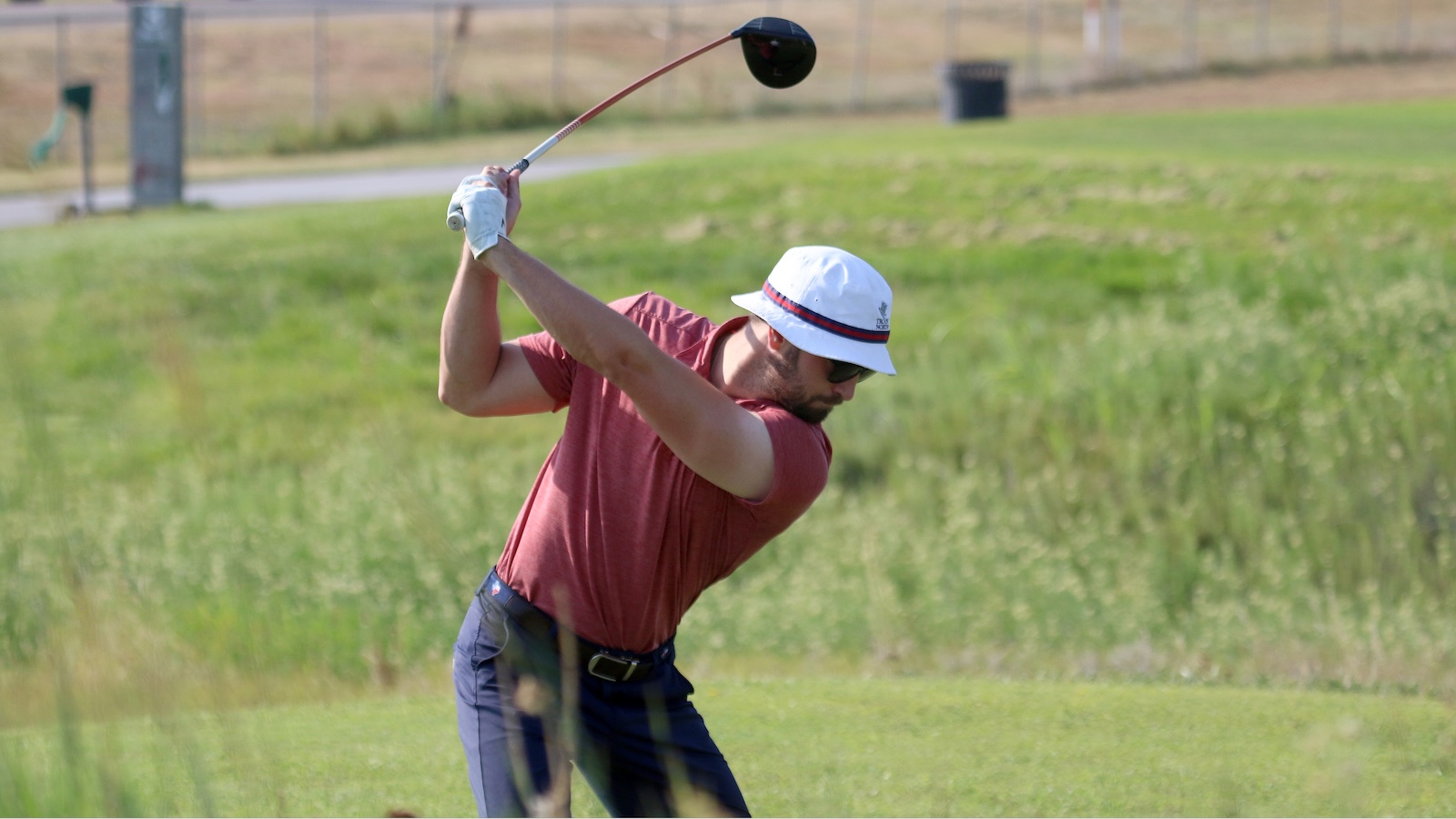 Golfer in a red shirt and white hat mid-swing on a grassy golf course field.