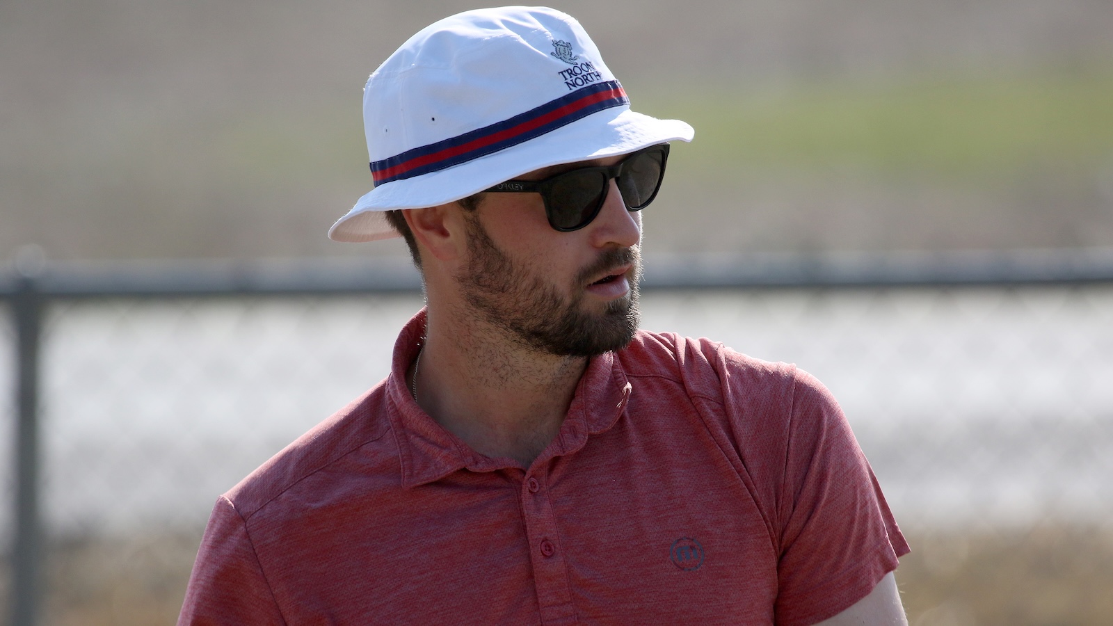 Man in a red polo shirt wearing dark sunglasses and a white bucket hat, looking to the side outdoors near a chain-link fence