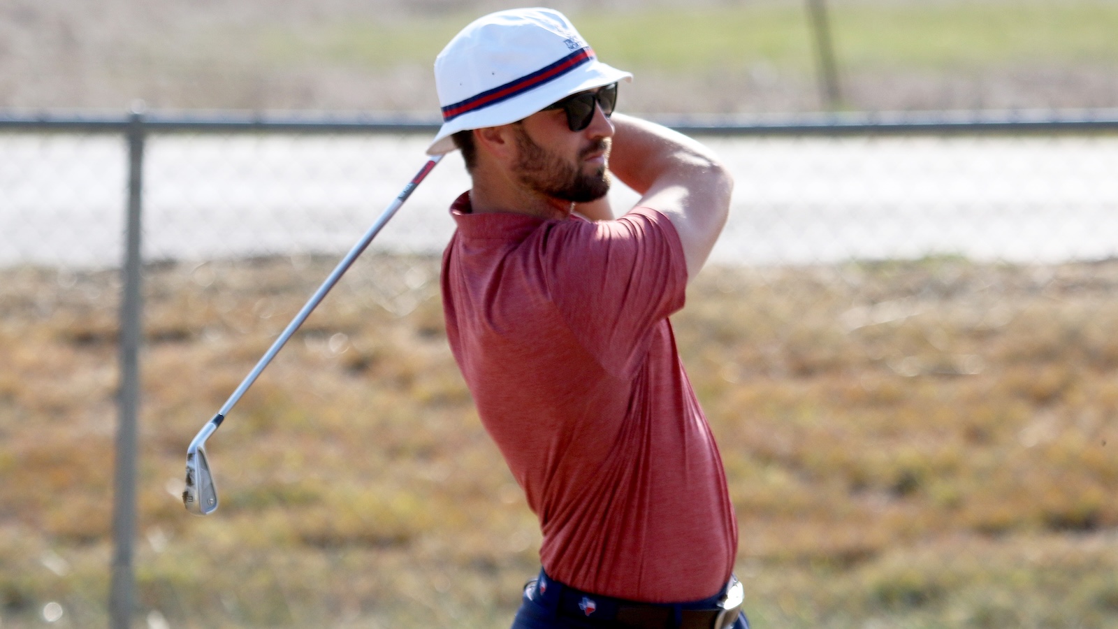 Man in a red polo shirt and white bucket hat swinging a golf club outdoors, wearing sunglasses with a fence and open field in the background