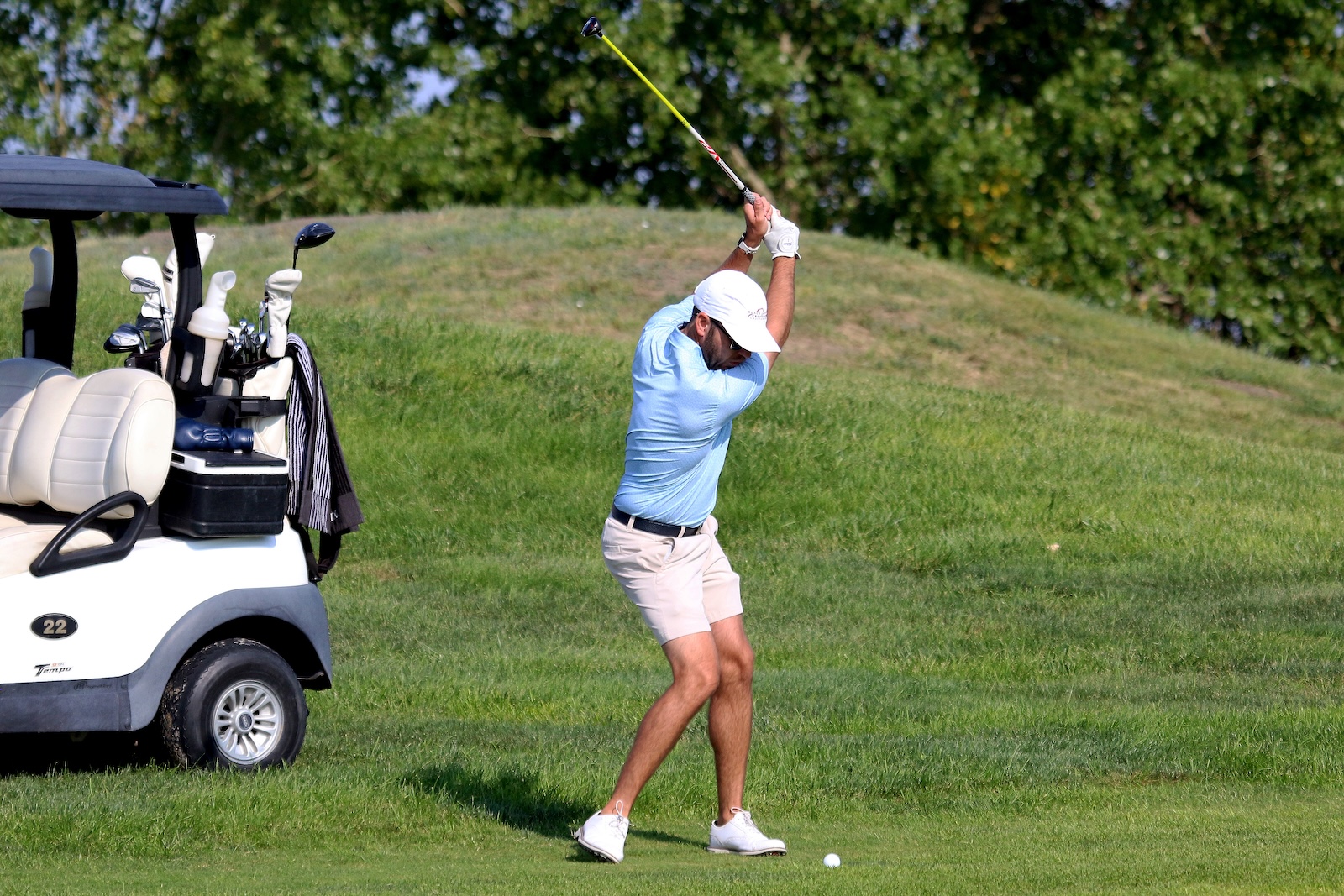 Golfer in light blue polo and beige shorts swings a golf club on a sunny course with a white golf cart and bags in the background.