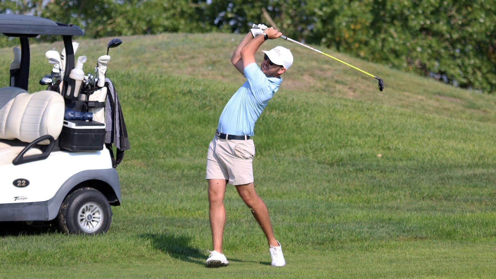 Golfer in a light blue shirt and beige shorts mid-swing on a grassy fairway beside a white golf cart with clubs.