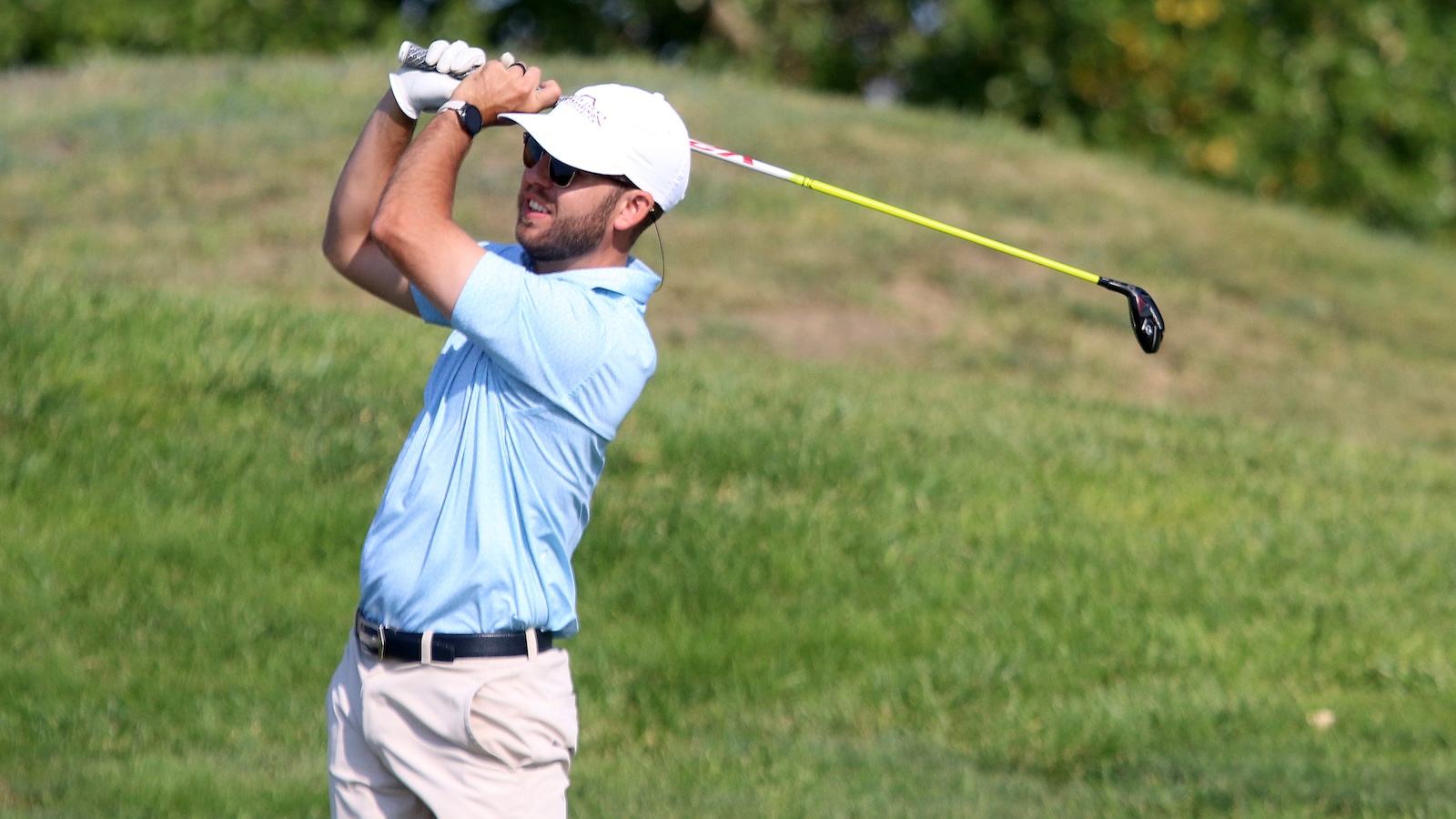 Golfer in a light blue polo and white cap mid-swing on a grassy golf course.