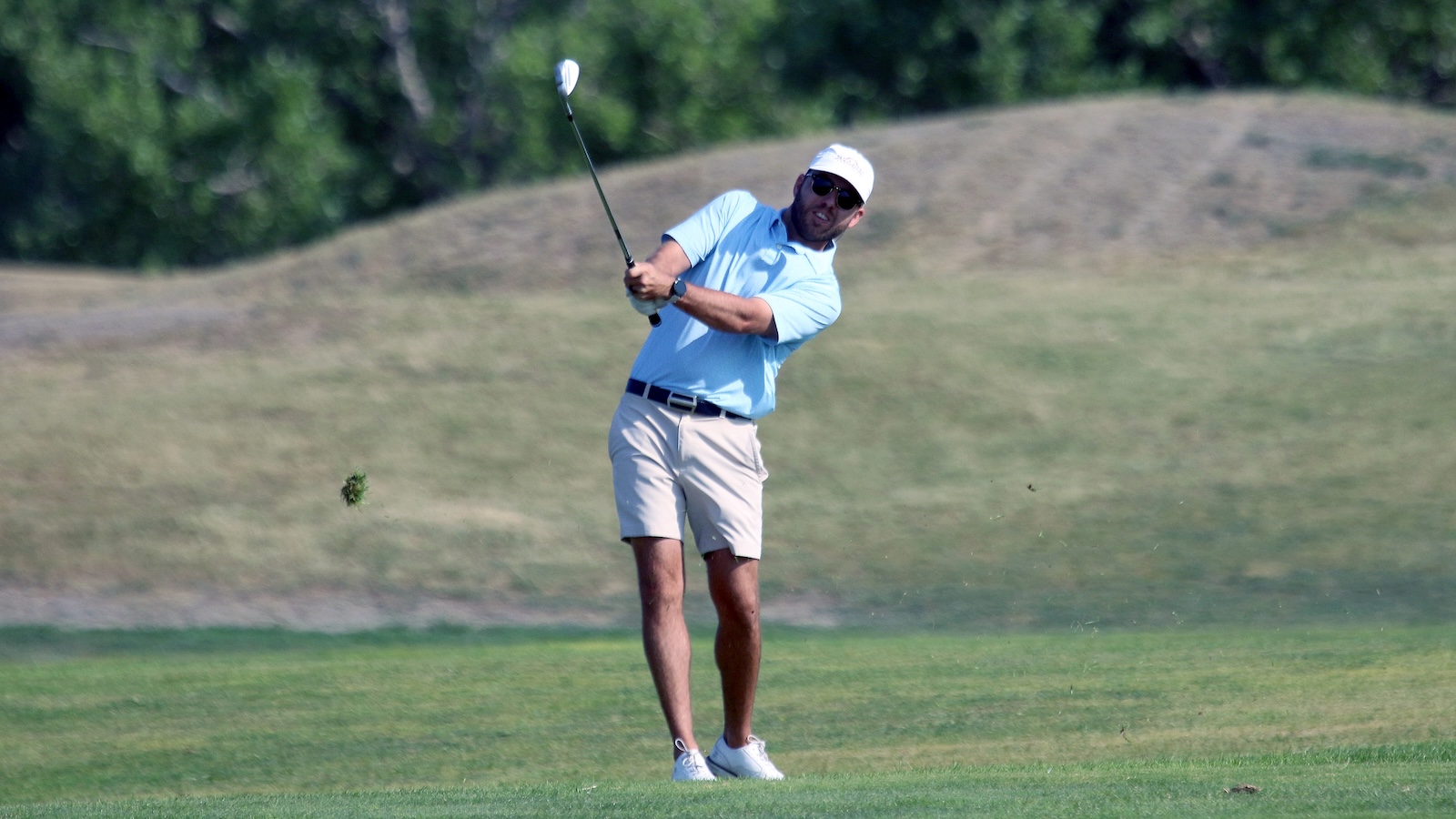 Man in a light blue polo and white cap mid-swing on a golf course, wearing sunglasses and white shoes.